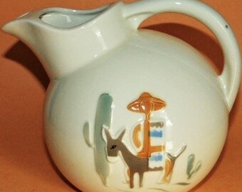 May include: Cream-colored ceramic pitcher with a rounded body and a handle. The pitcher features a Southwestern design with a donkey carrying a load, a cactus, and other desert-themed elements. The spout is designed for easy pouring.