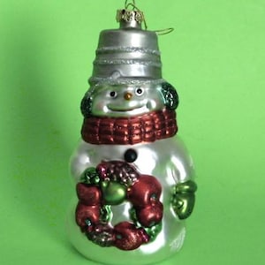 May include: A snowman Christmas ornament with a silver hat and body, a red scarf, and a green and red wreath. The ornament has a smiling face and a black button. The ornament is made of glass.