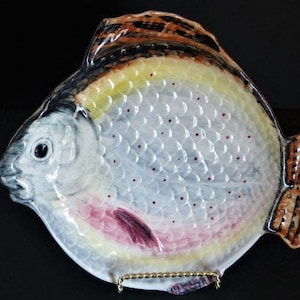 May include: A ceramic fish-shaped serving dish with a detailed, hand-painted design. The dish features a white body with a scale pattern, yellow and brown accents, and a red-pink underbelly. The tail and fin are brown. The dish is supported by a gold-colored metal stand.