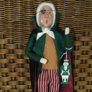 May include: A vintage-style doll dressed as a vampire with a white wig, green jacket, and red and white striped pants. The doll has a black eye patch and is holding a small toy soldier.