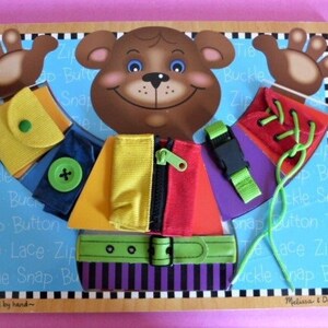 May include: A wooden toy with a brown bear face and a colorful outfit with different fasteners. The outfit has a yellow shirt with a zipper, a red vest with a snap button, a purple sleeve with a buckle, and a red shoe with laces. The toy is labeled "crafted by hand" and "Melissa & Doug".