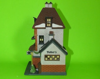 May include: A white and brown ceramic building with a sign that reads "Potter's". The building has a brown roof with a chimney and a small tree in front of it.