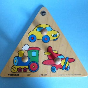 May include: A wooden triangle puzzle with three pieces: a yellow car, a red airplane, and a blue train. The puzzle is labeled "Vehicles" and "12+ MOS".