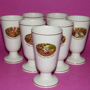 May include: Set of seven white ceramic coffee cups with gold trim and illustrations of coffee beans and coffee production. Each cup has a different label with the name of a coffee variety, including "Arabica" and "Robusta".