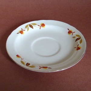May include: A white saucer with a gold rim and a pattern of brown leaves and orange flowers.