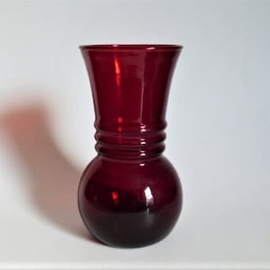 May include: A deep red glass vase with a rounded base and a flared opening. The vase has a textured design around the middle.