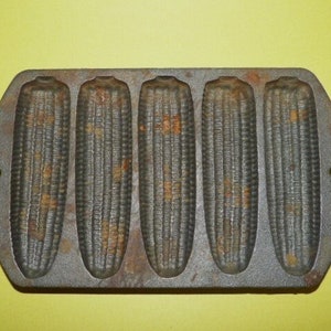 May include: A vintage cast iron cornbread pan with five individual molds shaped like corn cobs. The pan is rusty and has a dark gray color.