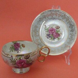 May include: A vintage teacup and saucer set. The cup and saucer are white with a pearlescent sheen, featuring pink rose floral designs and gold trim. The cup has a gold handle. The saucer is supported by a clear plastic stand.