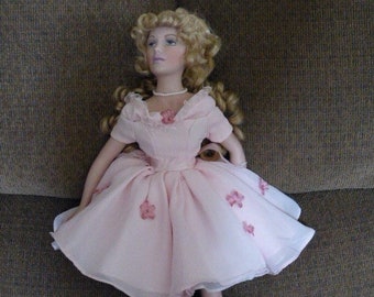 May include: A porcelain doll with blonde hair wearing a pink dress with floral accents. The doll is holding a small stuffed animal and is sitting on a brown couch.