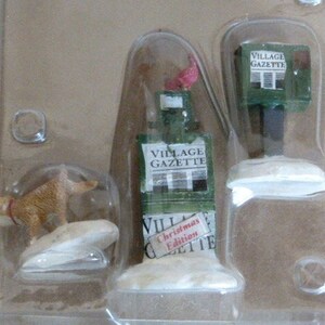May include: A set of three miniature Christmas village figurines. The figurines are a brown dog, a green newspaper stand with the text "Village Gazette" and a green newspaper stand with the text "Village Gazette Christmas Edition".