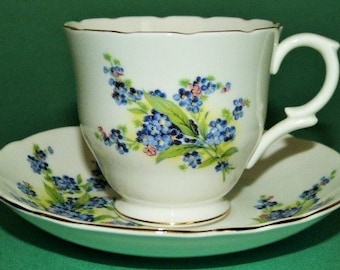 May include: A white teacup and saucer with a floral pattern of blue forget-me-nots and green leaves. The cup has a scalloped rim and a gold trim.
