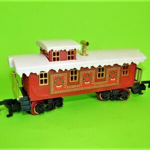 May include: A red and white toy train car with a snowy roof and gold accents. The car has windows and a wreath design on the side.