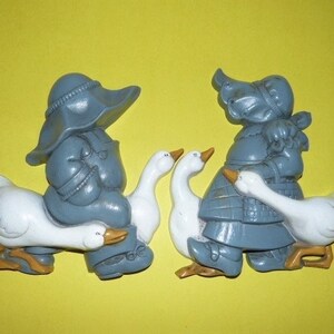 May include: Two gray figurines of people riding white geese. The people are wearing hats and long coats. The geese are facing forward with their wings outstretched.