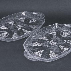 May include: Two clear glass serving trays with a grape and vine design. Each oval-shaped tray has a scalloped edge and a small handle. The grape clusters are white, and the trays are on a dark background.