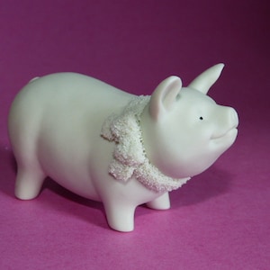 May include: White ceramic pig figurine with a white lace collar. The pig is standing on a pink surface.