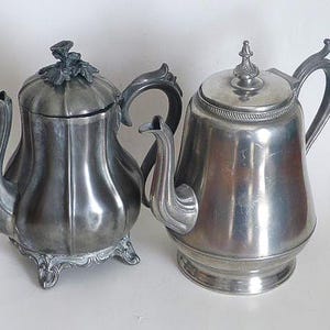 May include: Two antique silver teapots. One teapot has a lid with a floral design and ornate legs. The other teapot has a simple design with a decorative finial on the lid. Both teapots have curved spouts and handles.