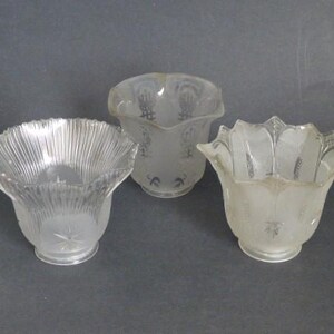 May include: Three vintage glass lamp shades. The shades have a clear, frosted, and etched design. The shade on the left has a ribbed texture, while the other two have floral patterns. The shades are in varying shapes and sizes.