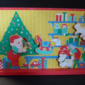 May include: A wooden Christmas-themed play set with a red base and a yellow background. The set features a green Christmas tree with ornaments, a red-suited elf holding a present, a rocking horse, a dog, a cat, a teddy bear, and a child holding a gift. The set also includes a shelf with presents and the word "Cookies" on a jar.