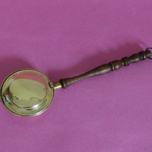 May include: A vintage brass warmer with a wooden handle and a small ring at the end. The round brass warmer has a lid and is set against a purple background. The handle is dark brown and has a carved design.