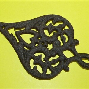 May include: A black cast iron heart-shaped trivet with a decorative, intricate design. The trivet has a loop on the top for hanging.