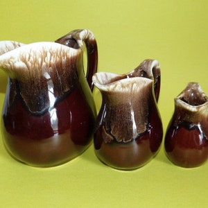 May include: Three brown ceramic pitchers with white glaze. The largest pitcher has a handle and a wide mouth. The two smaller pitchers have a smaller mouth and no handle.