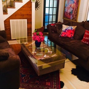 Könnte beinhalten: Ein Couchtisch mit Glasplatte, einem Holzrahmen und einem unteren Regal. Der Tisch hat einen rustikalen Look und ist mit einem Blumenarrangement und verschiedenen Gegenständen dekoriert, darunter Postkarten und Fotos. Der Tisch steht auf einem roten und schwarzen Teppich in einem Wohnzimmer mit einem braunen Sofa und einem weißen Kuhfellteppich.