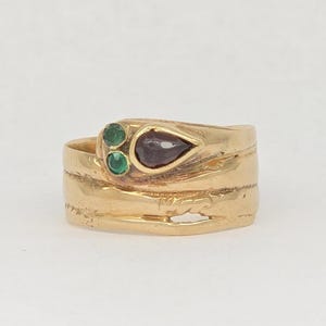 May include: A gold ring featuring a textured, wrap-around band design. The head of the ring is adorned with two small, round green stones and one larger, pear-shaped dark red stone, all set within the gold metal surface.