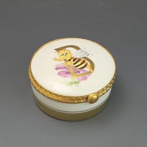 May include: A round white porcelain trinket box with a gold metal rim and hinge. The lid features a hand-painted illustration of a yellow and black bee resting on a large letter D, positioned above a small cluster of pink and green flowers.