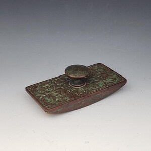 Pode incluir: Um tinteiro retangular antigo de bronze com um botão circular. O tinteiro tem um acabamento pátina e é decorado com padrões circulares em relevo. Este artigo de escritório vintage mede aproximadamente 10 cm de comprimento.