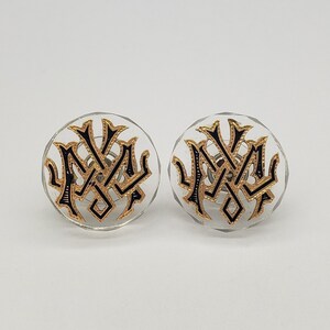 May include: A pair of clear acrylic earrings with a black and gold New York City logo design.