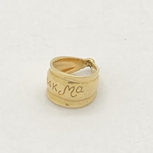 Puede incluir: Un pequeño anillo dorado con la inscripción "mk.ma" grabada en su superficie. El anillo tiene forma cilíndrica con una textura ligeramente irregular, lo que sugiere que puede ser hecho a mano. El fondo es una superficie lisa de color blanquecino.