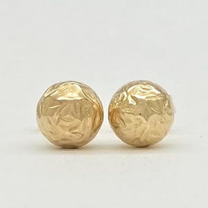 May include: A pair of spherical stud earrings crafted from polished yellow gold. Each earring features a textured, hammered surface with irregular indentations that catch the light, creating a multifaceted appearance on the round, ball-shaped design.