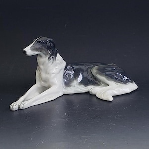May include: A porcelain figurine of a black and white dog lying down. The dog has a long, slender body and a long, pointed snout.