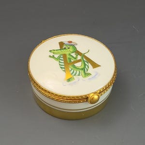 May include: A small, round, white porcelain trinket box with a gold-colored metal rim and hinge. The lid features a green alligator illustration wearing a white sailor hat, positioned in front of a large gold letter A, with a gold base.