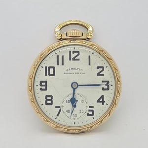 May include: A gold-coloured Hamilton Railway Special pocket watch. The watch has a white face with black numerals and a small seconds dial. The hands are blue. The watch case has an ornate design around the edge.