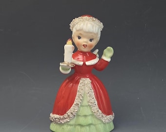 May include: A ceramic figurine of a person with white hair wearing a red coat with white trim and a light green skirt. The figure holds a white candle with a yellow flame in one hand and has glittery accents on the hair, collar, and hem.