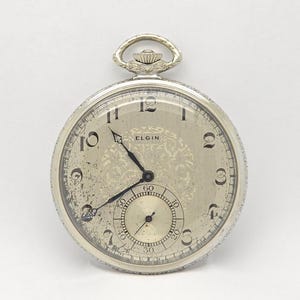 May include: A silver-toned Elgin pocket watch with a round face, black Arabic numerals, and ornate filigree detailing in the centre. The watch features a sub-dial for seconds at the bottom, black hands, and a decorative winding crown at the top.