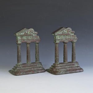 May include: A pair of bookends featuring a classical architectural temple design with three fluted columns, a stepped base, and a triangular pediment. The metal objects have a weathered, dark bronze finish with green patina accents throughout.