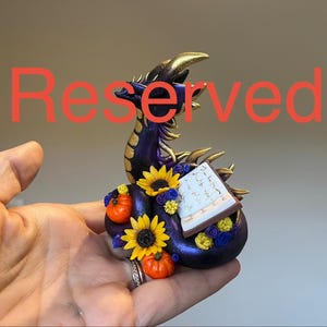 May include: A small purple dragon figurine with gold accents, featuring a coiled tail, two orange pumpkins, yellow sunflowers, small blue and yellow flowers, and an open book with visible text, all overlaid with the red text Reserved.