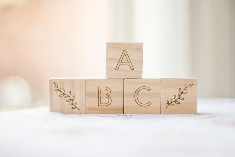 Pode incluir: Blocos de madeira com as letras A, B e C, e um desenho de folhas nas laterais. Os blocos est&atilde;o empilhados em forma de pir&acirc;mide.