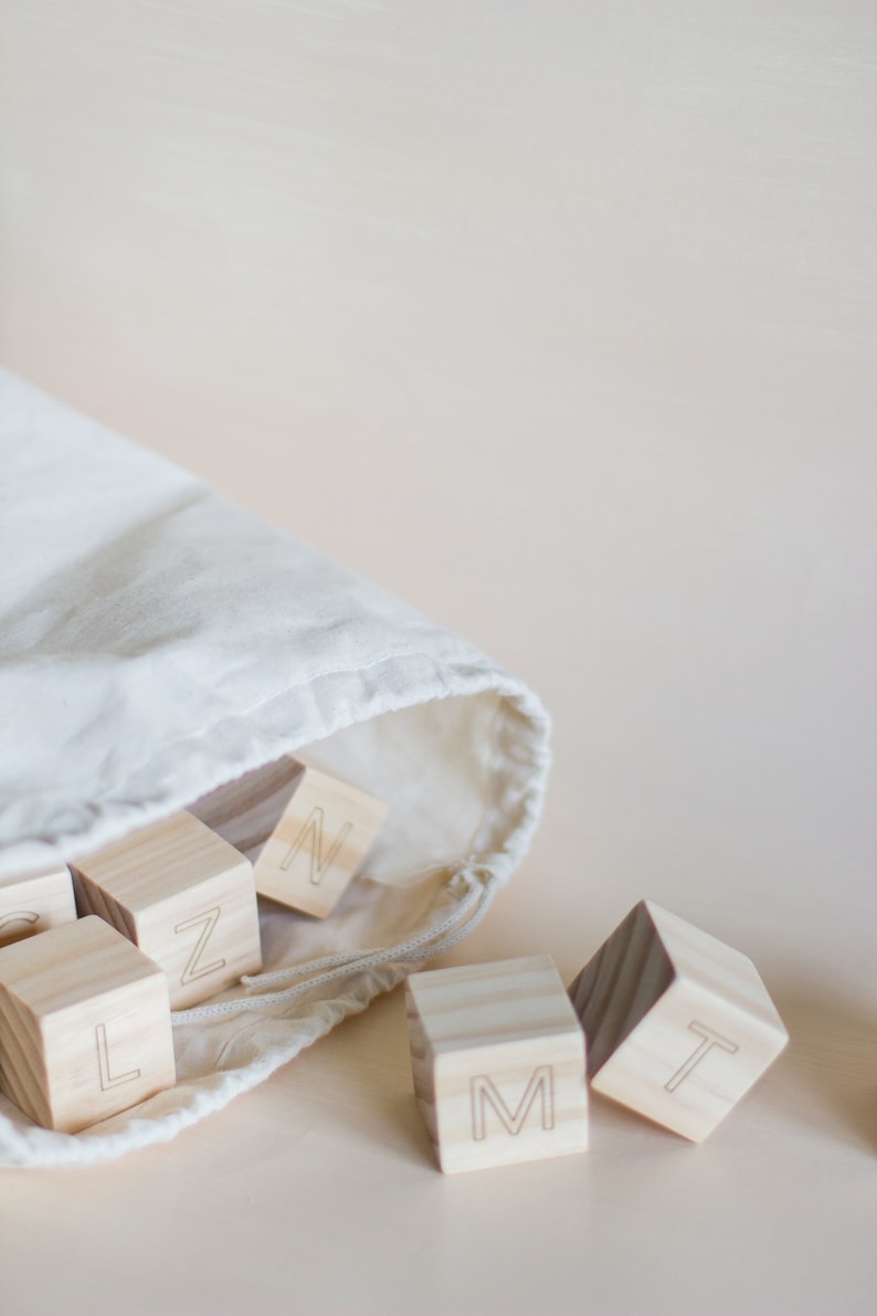 May include: A white canvas bag with wooden blocks that have letters on them. The letters are L, Z, N, M, and T. The blocks are spilling out of the bag.