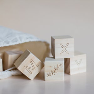 Pode incluir: Blocos de alfabeto de madeira com as letras M, X, Y e um desenho de folha. Os blocos est&atilde;o em uma bolsa de tecido branca.