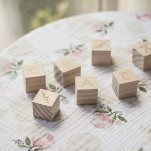 Pode incluir: Blocos de alfabeto de madeira com as letras A, B, C, E, Y e X, e um bloco com um desenho de folha. Os blocos est&atilde;o sobre um tecido floral branco e rosa.