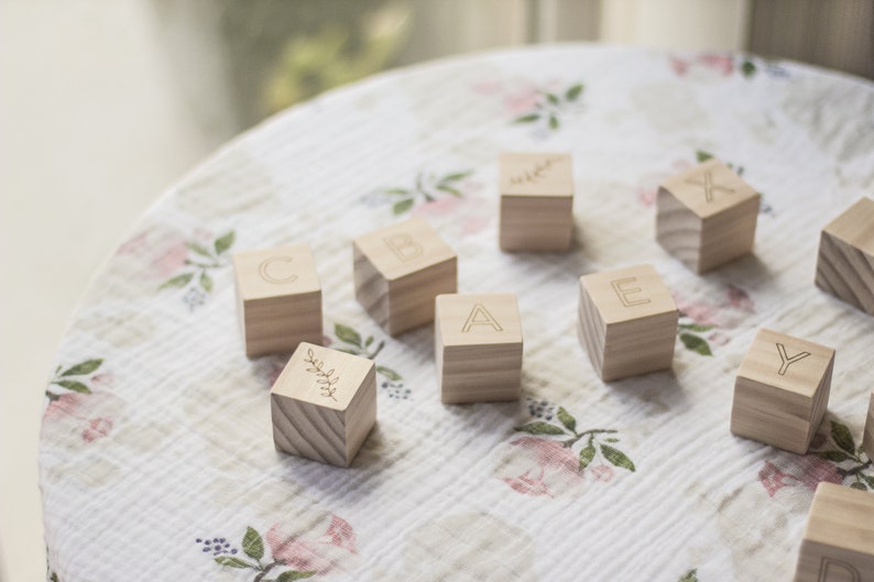 Pode incluir: Blocos de alfabeto de madeira com as letras A, B, C, E, Y e X, e um bloco com um desenho de folha. Os blocos est&atilde;o sobre um tecido floral branco e rosa.
