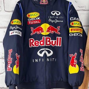 May include: A navy blue Red Bull racing jacket with various sponsor logos, including Infiniti and Pepe Jeans. The jacket has a full zip, a stand-up collar, and Red Bull branding on the sleeves and chest.