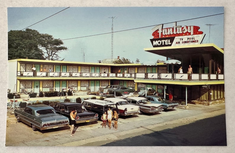 May include: Vintage postcard featuring the Fantasy Motel with a retro sign. The two-story building has a pool and is surrounded by classic cars. The sign reads "Fantasy Motel TV Pool Air Conditioned". People are standing on the balcony and in front of the motel.
