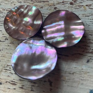 Op de afbeelding: Drie iriserende, ronde knopen met een parelmoer effect. De knopen hebben een donkerbruine basiskleur met glinsterende highlights van roze, paars en groen. De knopen zijn gerangschikt op een lichtgekleurd, getextureerd oppervlak.
