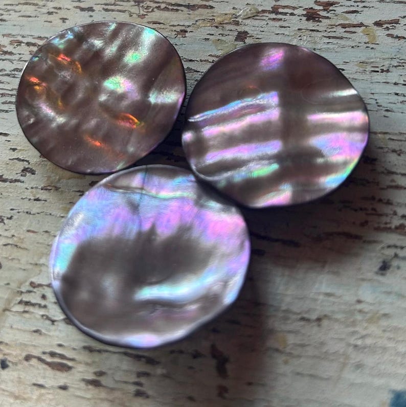 Op de afbeelding: Drie iriserende, ronde knopen met een parelmoer effect. De knopen hebben een donkerbruine basiskleur met glinsterende highlights van roze, paars en groen. De knopen zijn gerangschikt op een lichtgekleurd, getextureerd oppervlak.