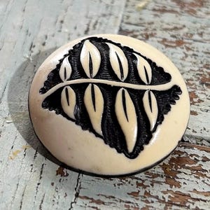 May include: A round, off-white button with a black leaf design. The button has a dark rim and a detailed, carved leaf pattern. The button is likely made of a hard material.
