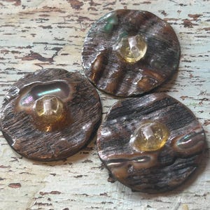 Op de afbeelding: Drie ronde, donkerbruine knopen met een getextureerde, houtachtige uitstraling. Elke knoop heeft een centrale, heldere, amberkleurige edelsteen. De knopen hebben iriserende highlights en zijn gerangschikt op een verweerd, lichtgekleurd houten oppervlak.